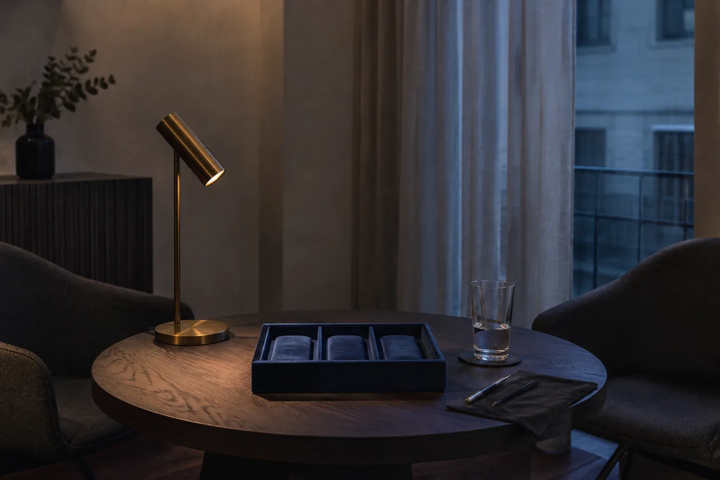 The viewing room: a round oak table with an empty navy tray for three watches, two armchairs, a brass lamp, blue hour in the window